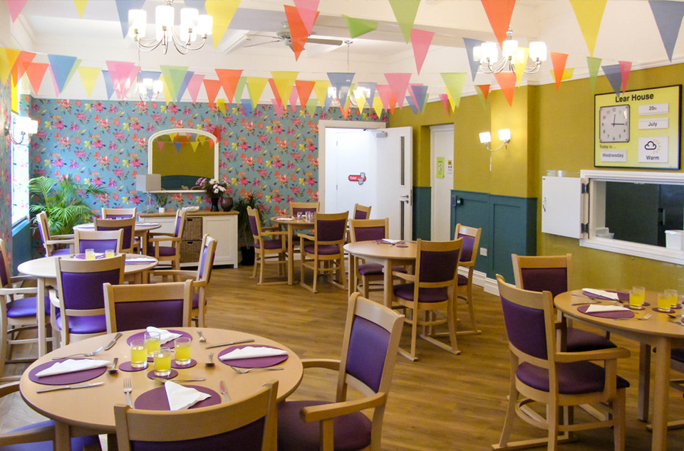 Brightly decorated dining area with round tables, purple chairs, colourful bunting, and floral wallpaper. Tables are set with glasses of orange juice and place settings.