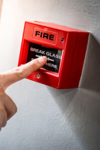 A finger pressing the break glass button on a red fire alarm box mounted on a wall, ensuring safety in a Wirral care home.