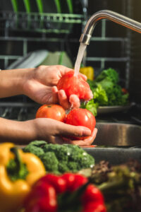 In the kitchen of a residential care home, a person washes three tomatoes under the tap, with colourful peppers and fresh broccoli in the foreground.