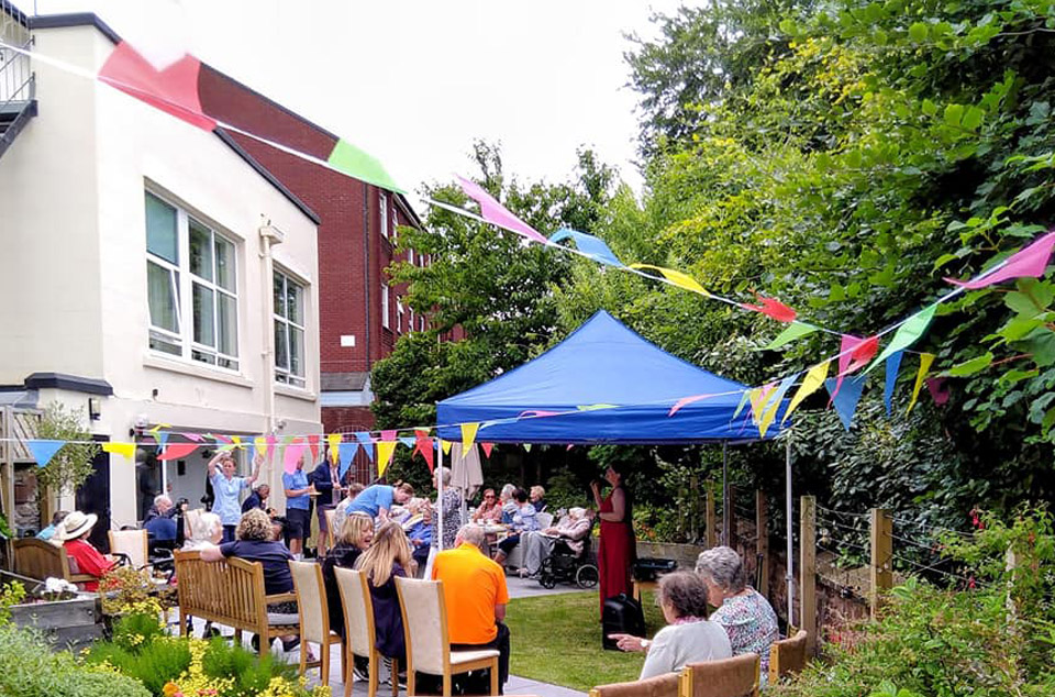 A group gathers in a garden under colourful bunting and a blue canopy at the Wirral care home. Some are seated while others stand, enjoying the day with a two-storey building and trees in the background.