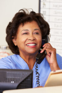 Seated in front of a computer screen, the person in a blue shirt smiles warmly whilst speaking on a corded phone, holding a folder. It’s another day at Abbeyfield Wirral Care Home, where the focus is on creating meaningful connections.