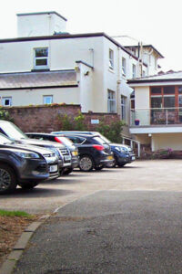 A row of parked cars is situated in front of a multi-storey building, identified as a residential care home, with a welcoming lower entrance and gleaming windows.