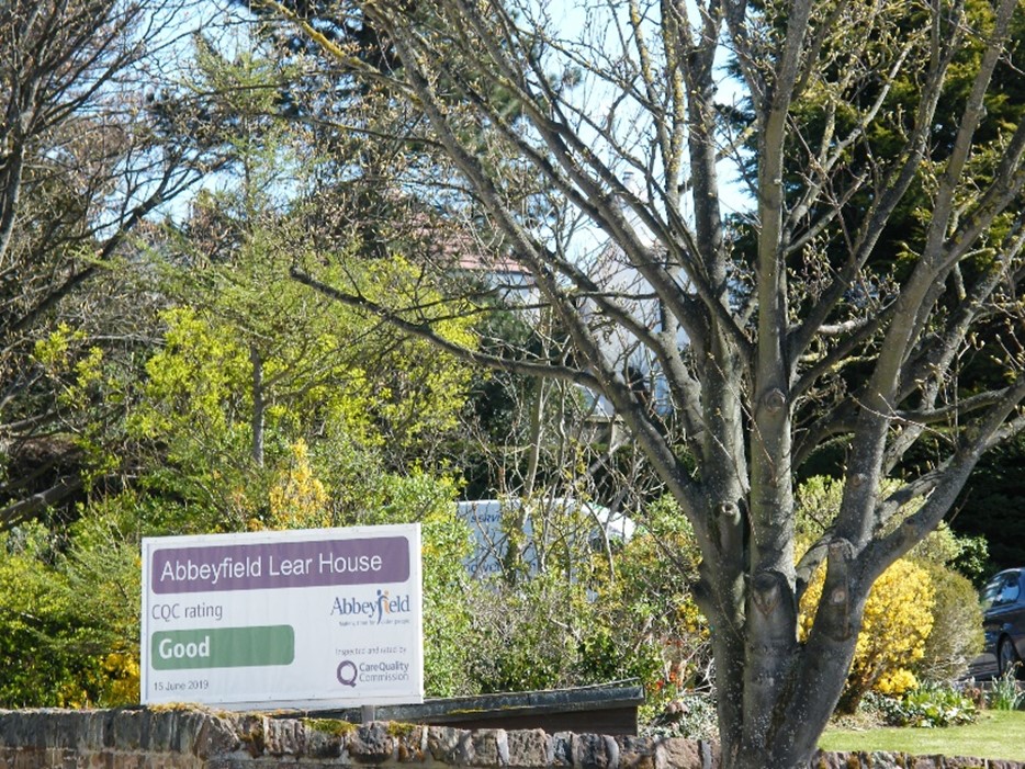 The sign for Abbeyfield Lear House, a residential care home with a Good CQC rating, stands gracefully among trees and bushes.