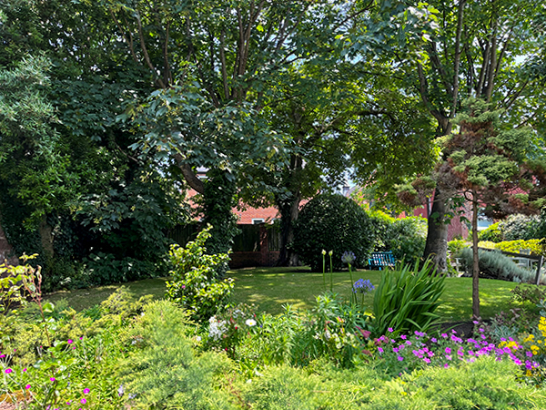 A lush garden scene surrounds the Wirral care home, where various trees and flowering plants thrive. Sunlight filters through the leaves, casting dappled shadows on the grass, creating a serene retreat for residents in supported care.