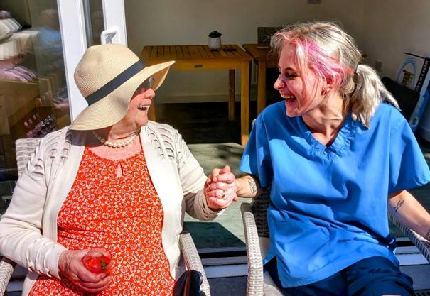 An elderly woman in a sunhat holds hands and smiles with a young woman in blue scrubs, sitting outdoors at Abbeyfields tranquil residential care home.