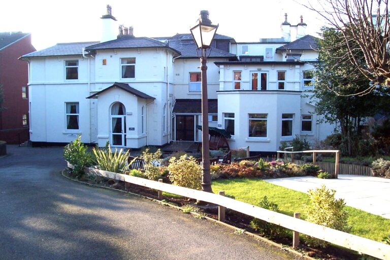 A large, white house with multiple storeys and a welcoming garden at the front serves as an inviting Wirral care home. There’s a lamp post and path leading gracefully to the entrance, embodying the warmth and community spirit of Abbeyfield.