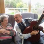 An elderly woman smiles at a man in a suit playing a guitar as they sit together on armchairs in the cosy Abbeyfield care home. Behind them, a window offers a serene outdoor view, enhancing their tranquil respite moment.