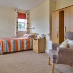 A tidy bedroom in a Wirral care home features a single bed with orange striped bedding, a wooden wardrobe, and a small side table with a teapot. A chest of drawers and an armchair near the window with curtains complete the comforting setup for assisted living.