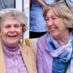 Two older women with grey hair, wearing spectacles and light-coloured clothes, are smiling warmly as they sit together indoors at a cosy Abbeyfield residential care home.