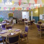 A colourful dining room in the Wirral care home features round tables laid with purple serviettes and glasses, while bright bunting hangs from the ceiling. The walls are adorned with floral and mustard-yellow accents, creating a welcoming space for residents enjoying respite care.