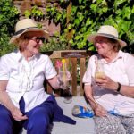 Two elderly women wearing hats sit outdoors on a bench at Abbeyfield Hoylake & West Kirby, smiling and holding drinks with straws and umbrellas. One has a knitting project beside her. A plaque on the bench is visible, adding a touch of charm to this tranquil scene.