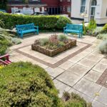 A small paved courtyard at the residential care home features two blue benches and one red bench. A central square planter is flanked by a variety of shrubs and greenery along the edges, providing a peaceful retreat for residents of the abbeyfield community.