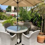 On the patio of our assisted care home, youll find a round glass table with five wicker chairs shaded by a large beige umbrella. Surrounded by greenery and landscaping, this tranquil spot under the clear blue sky provides a peaceful retreat.