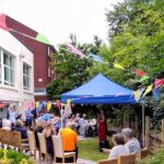 A group of people gather in a garden under a blue canopy, with colourful bunting overhead at the Wirral Care Home. Some are seated on chairs, while others stand or chat near a brick building, enjoying the lively atmosphere and camaraderie that comes with assisted care living.