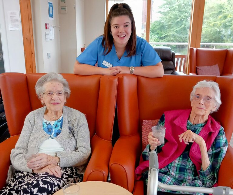 Two elderly women sit on orange chairs, each holding a drink, while a smiling healthcare worker in uniform leans behind them indoors at Abbeyfield Hoylake & West Kirby.