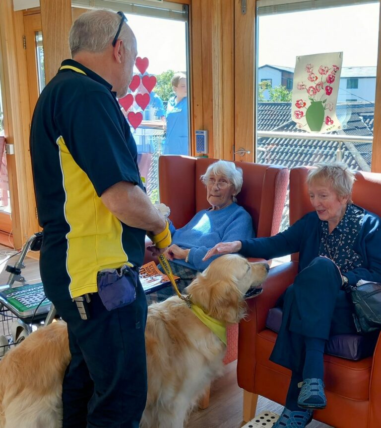 Two elderly women sit in chairs, stroking a golden retriever held by a man in a black and yellow shirt at the cosy Abbeyfield care home. The room has wooden walls and large windows with a sunny view, creating a warm and inviting atmosphere.