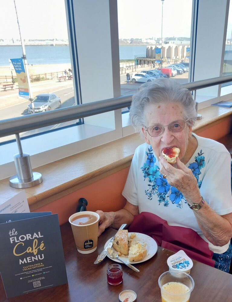 An elderly woman, taking a break from her seaside respite care on the Wirral, enjoys a meal at a café. She savours a scone with her hot drink, while another scone awaits on the table, perfectly complementing the scenic view outside the window.