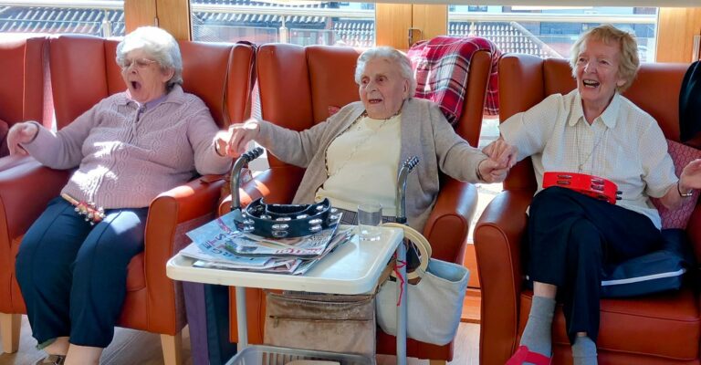 Three elderly women in a Wirral care home sit in chairs, holding hands and playing musical instruments.