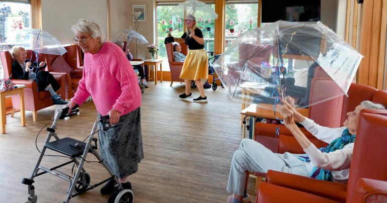 Residents of the care home gather in a cosy common room, one using a walking frame while others sit comfortably with see-through umbrellas. In the background, a carer stands holding an umbrella, ensuring everyone feels safe and sheltered.