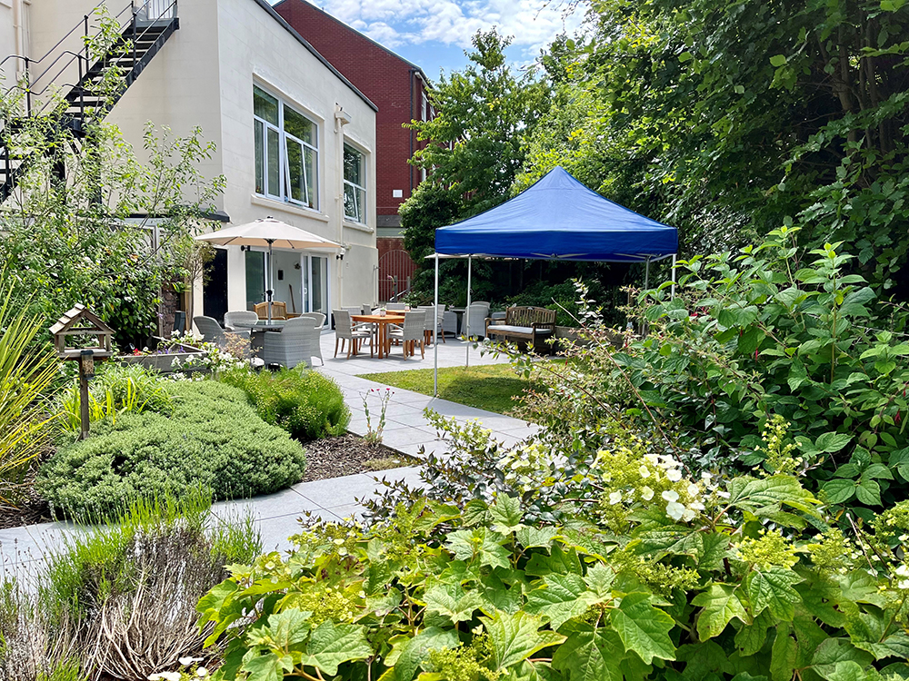 The garden of the assisted care home features a patio set under a parasol, complemented by a blue canopy. Lush greenery surrounds a charming bird box, while a white building with an external staircase stands gracefully in the background.