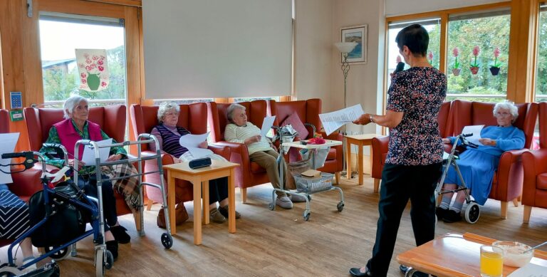 In a bright room with wooden floors and large windows, a group of elderly residents at the residential care home sit in armchairs, attentively listening to a woman reading from sheets of paper.