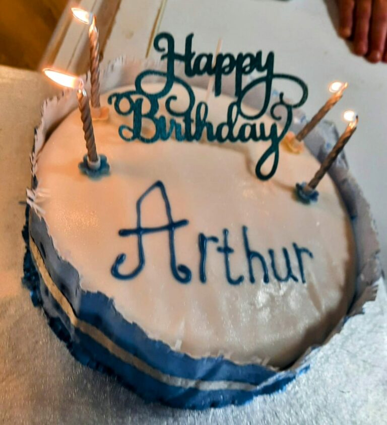 Round cake with blue-and-white icing and Happy Birthday Arthur written on top. Four lit candles adorn the cake, celebrating a special day at the Wirral care home. A pair of hands are partially visible in the background, adding a touch of warmth to this heartfelt celebration.