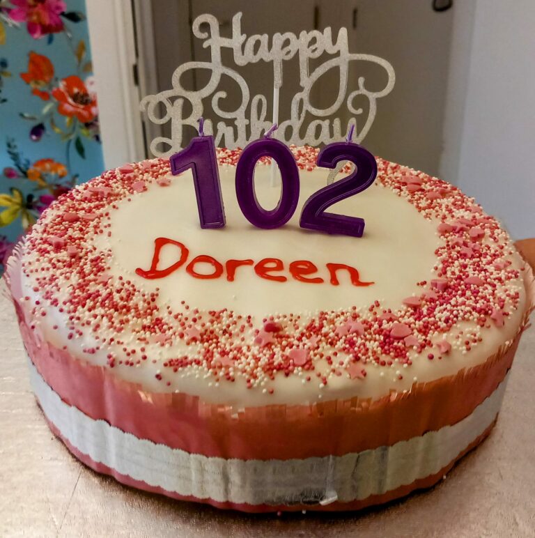 Birthday cake with white icing, pink sprinkles, and purple 102 candles sits on the table at Abbeyfield. A Happy Birthday topper and Doreen, written in red icing, celebrate another cherished year in our residential care home.