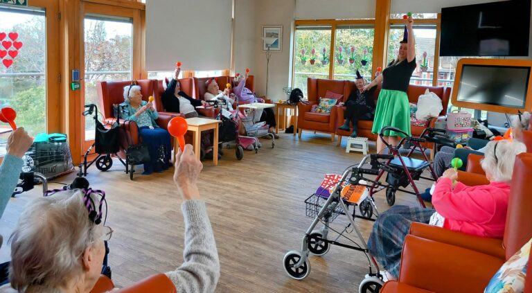 In a bright room adorned with hearts, a lively group of elderly residents at an assisted care home shake red maracas, guided by a spirited woman. Large windows flood the space with light, creating a joyful atmosphere.