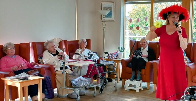 A woman in a red dress and hat sings into a microphone at a care home. Seated elderly residents listen attentively in the cosy room, where armchairs and a walking frame are visible.