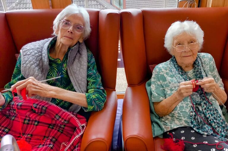 Two elderly women sit in armchairs, knitting with red wool, their white hair and glasses glinting softly in the cosy setting of a care home.