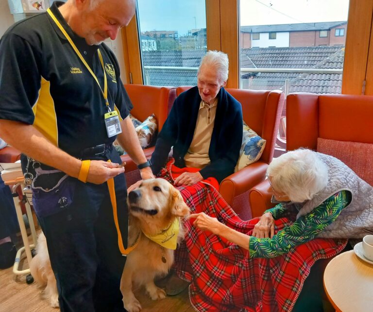 A man in a yellow and black shirt stands with a golden retriever at the Wirral Care Home. Two elderly residents sit on a sofa, stroking the dog, with a red tartan blanket over their laps, enjoying their respite care in this cosy Abbeyfield setting.