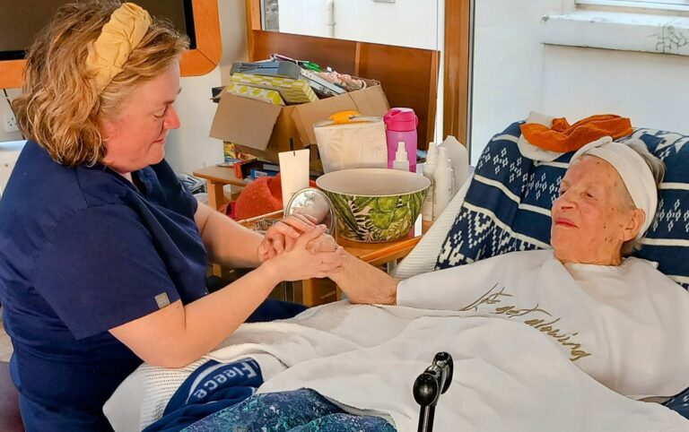 In a cosy assisted care home, a carer gently holds the hands of an elderly woman in bed, providing comfort and care amidst personal items scattered around.