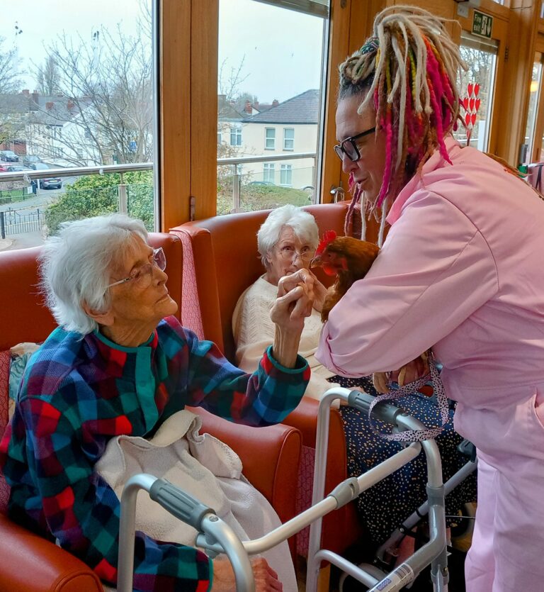 At Abbeyfield, a person with plaited hair in a pink outfit gently holds a small brown goat up to an elderly woman wrapped in a checked blanket, as another resident looks on in delight.