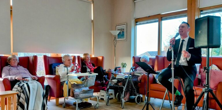 A man in a suit sings into a microphone, seated next to a music stand at the Wirral care home. Four elderly women listen, seated on chairs, as sunlight streams through large windows.