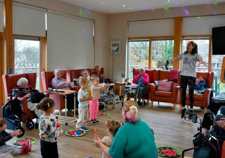 Children and elderly people are joyfully engaging in a group activity at the community room of an Abbeyfield care home, with colourful lights dancing on the ceiling.