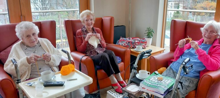 Three elderly women sit in armchairs, knitting with wool in their cosy Abbeyfield residential care home. A book titled Goodwood rests on a table alongside a cup and remote control, adding to the tranquil atmosphere of their communal living space.