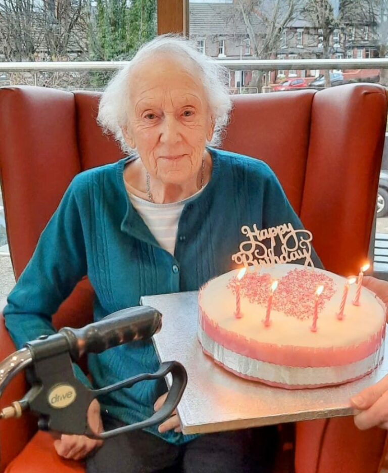 An elderly woman sitting in a chair at the care home holds a birthday cake with five lit candles and a Happy Birthday topper. A walking frame is visible in front of her.