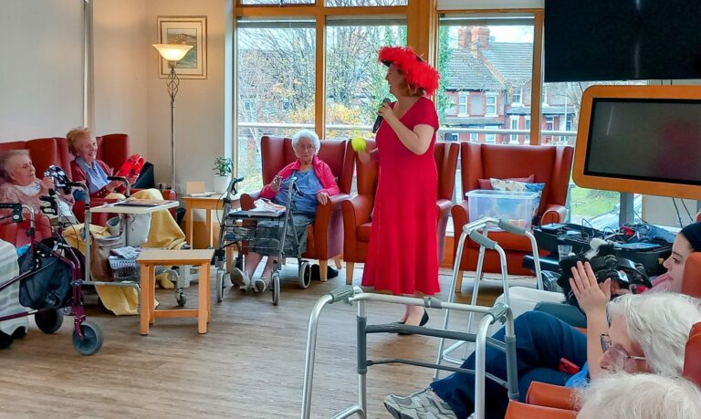 A woman in a red dress and hat performs with a microphone, captivating the seated elderly residents of the care home, surrounded by walking aids.