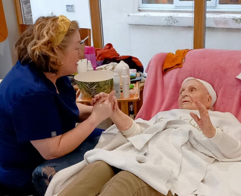 A woman in a blue uniform holds hands with an elderly person lying on a reclining chair, covered by a white blanket, in an Abbeyfield residential care home. Various personal care items are on a table nearby.