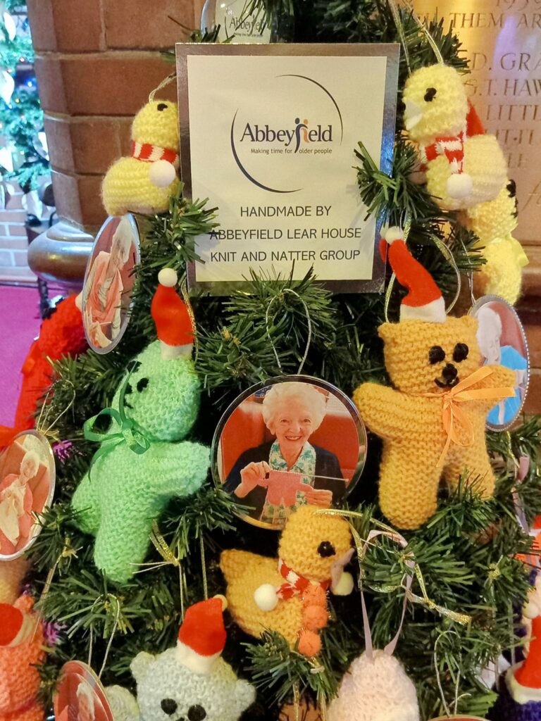 The Christmas tree, adorned with knitted teddy bear decorations and circular photos, boasts a sign that reads Abbeyfield, Handmade by Abbeyfield Lear House Knit and Natter Group, adding a festive touch to the Wirral care home.