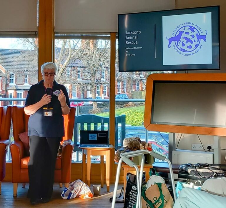 A woman delivers a presentation on Jacksons Animal Rescue in a room with a monitor displaying the logo and name, drawing parallels to the compassionate environment found in Abbeyfield residential care homes.