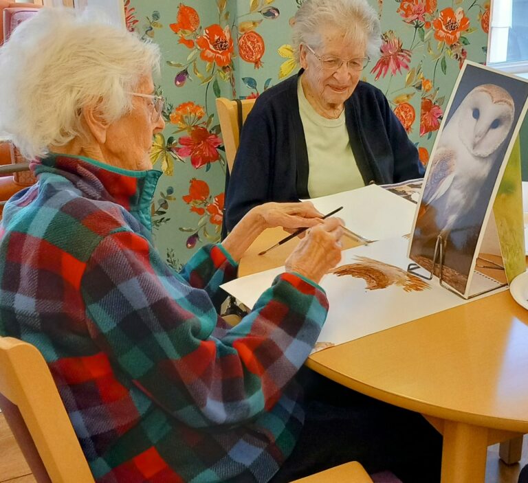 Two elderly women sit at a table in the Wirral care home, engaged in painting. One uses a reference image of an owl, while the floral-patterned curtain gently frames their creative moment.