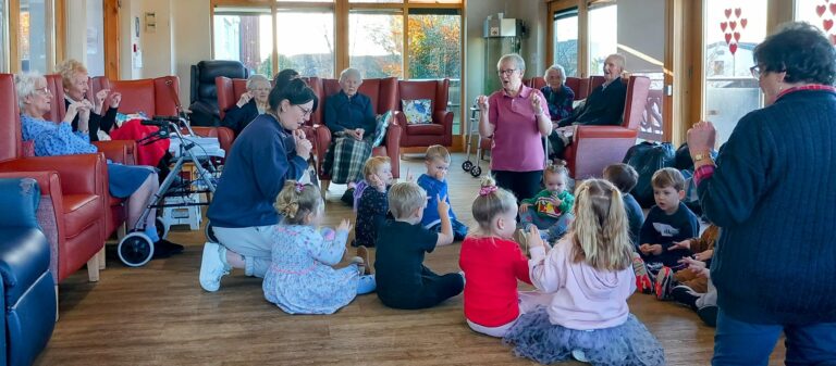 In an Abbeyfield assisted care home, children and adults gather in a room for an interactive activity. Two adults stand guiding the children, while others are seated, observing and participating. It’s a lively scene fostering connection across generations.