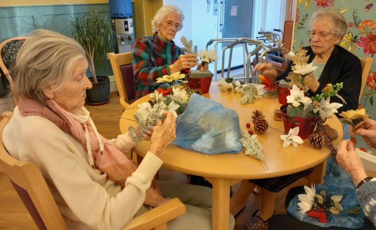 Four elderly women are seated at a table in the care home, creating beautiful floral arrangements with pinecones and artificial flowers.