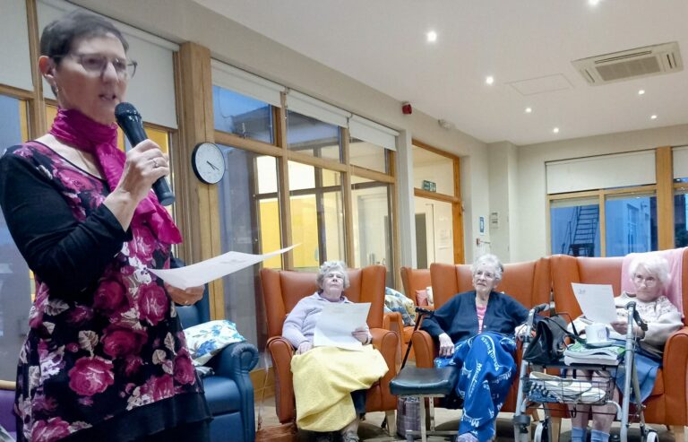 A woman with a microphone engages warmly with three seated elderly women in an Abbeyfield care home, sunlight streaming through large windows.