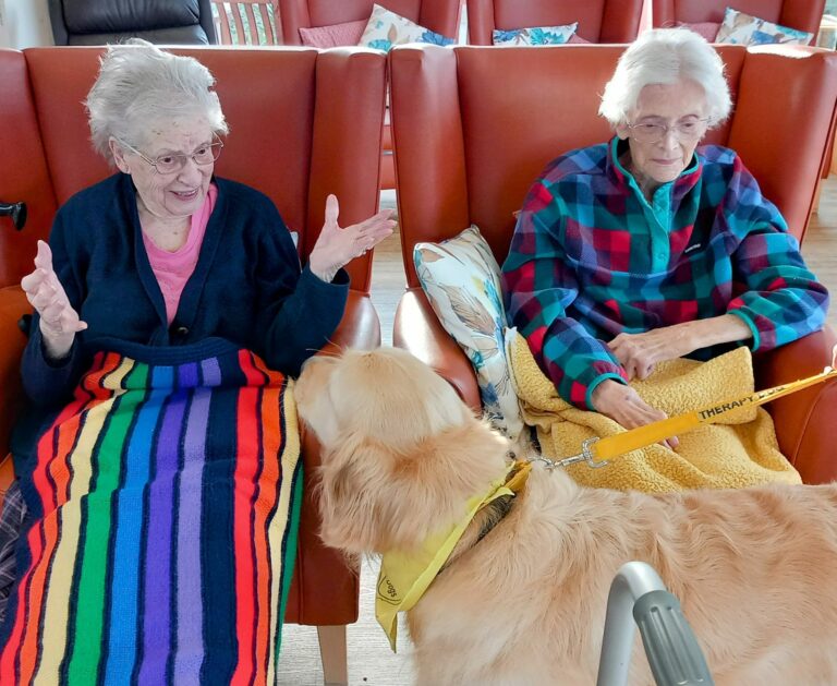 Two elderly women sit in armchairs, one with a colourful blanket, in the cosy setting of a care home. A golden retriever wearing a yellow therapy dog bandana stands warmly in front of them.