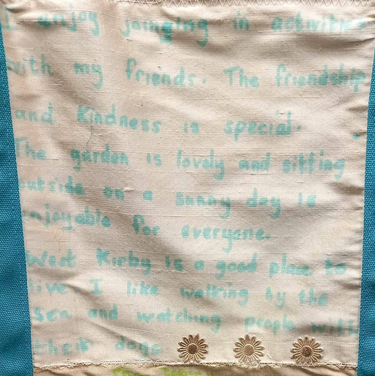 Fabric with handwritten text in blue ink, referencing friendship, kindness, enjoying time outdoors, and a place called West Kirby. Inspired by life at a Wirral care home, three daisy-like flowers are embroidered at the bottom.