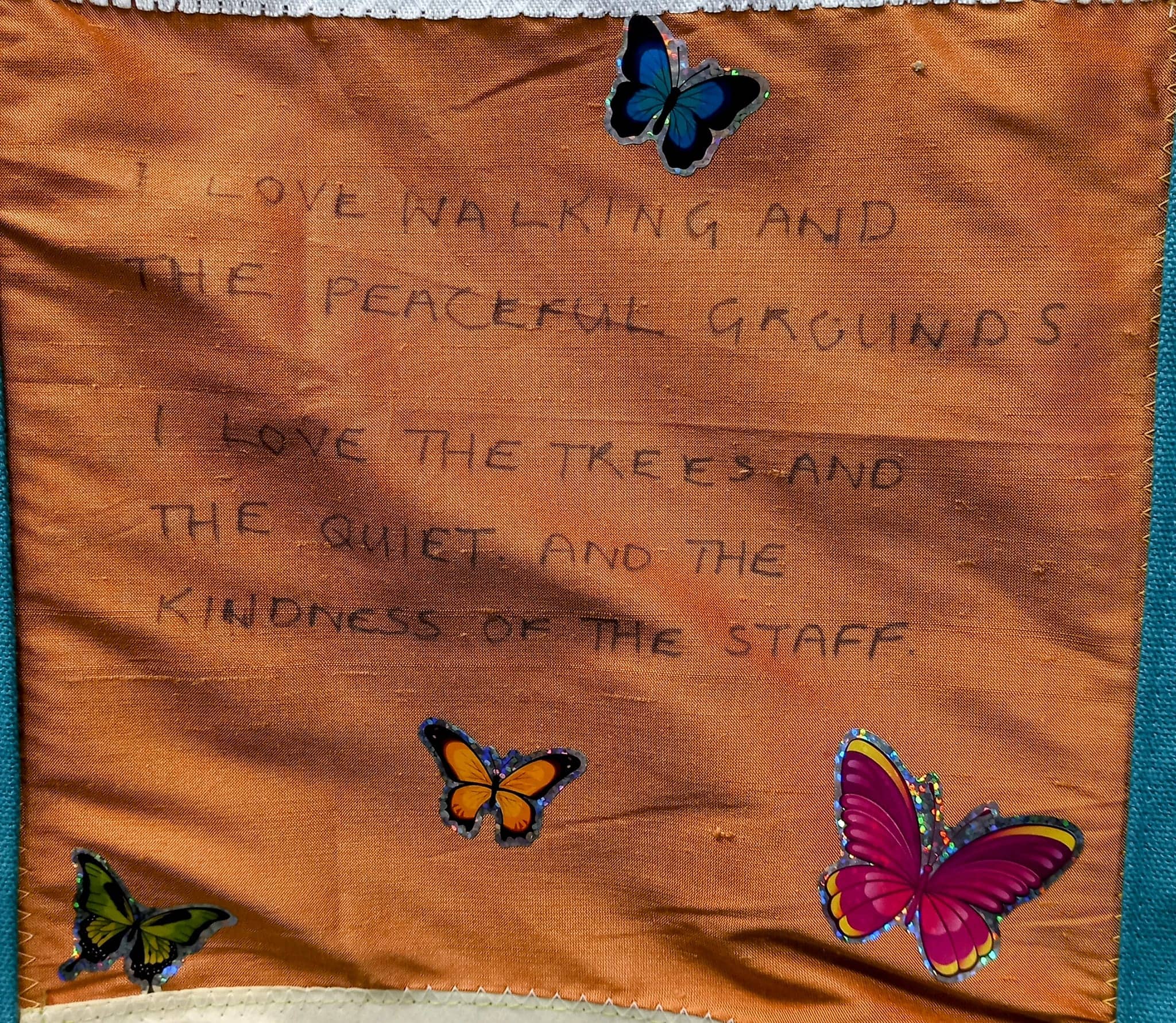 Handwritten text on fabric expresses appreciation for the peaceful grounds, trees, quiet, and staff kindness at the Wirral care home, beautifully decorated with colourful butterfly stickers.