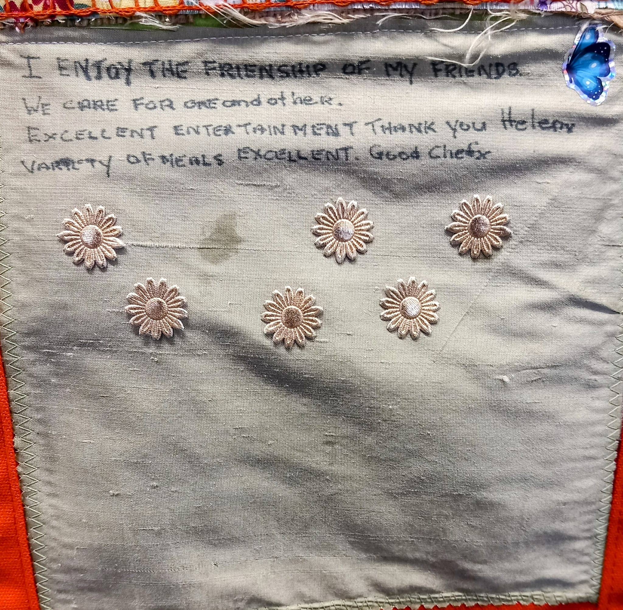 A handwritten note on fabric, mentioning friendship, entertainment, and meals at the Wirral care home, is beautifully adorned with embroidered flowers and a butterfly patch in the corner.