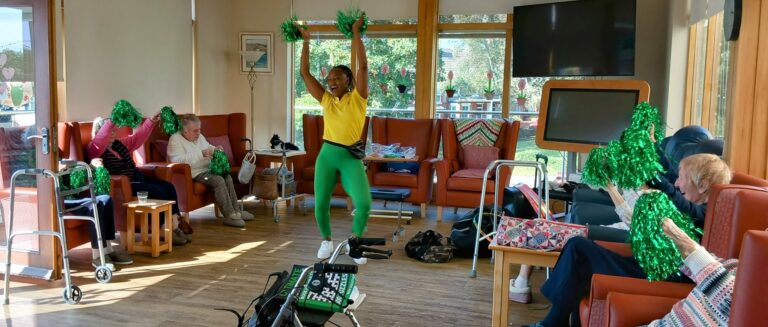 In an Abbeyfield residential care home, someone leads a lively cheerleading session with green pom-poms. Enthusiastic residents, seated in chairs, join in the fun and feel the vibrant energy of the moment.